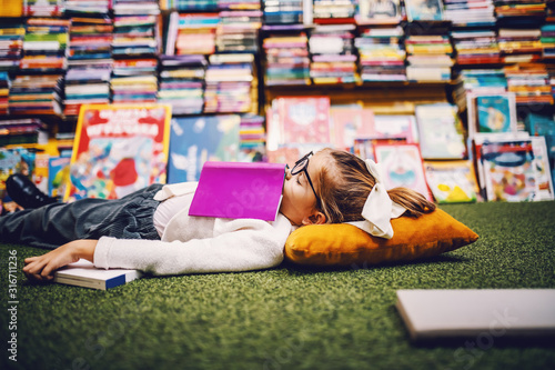 Charming caucasian little blond girl with ponytail lying on the floor in bookstore with book on chest and sleeping Tableau sur Toile