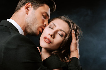 Wall Mural - handsome man kissing young woman with closed eyes on black with smoke