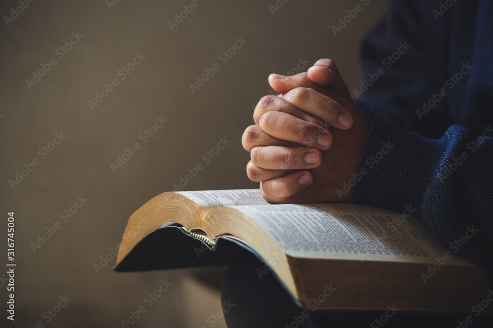 Hands folded in prayer on a Holy Bible in church concept for faith - obrazy, fototapety, plakaty 