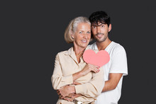 Young Man Embracing Senior Woman Holding Red Paper Heart Against Black Background