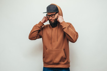 Wall Mural - Handsome hipster guy with beard wearing brown blank hoodie or hoody and black cap with space for your logo or design on white background. Mockup for print