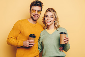 cheerful couple looking at camera while holding coffee to go on yellow background