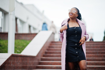 Wall Mural - Young stylish beautiful african american woman in street against stairs, wearing fashion outfit coat, eyeaglasses.