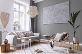 Fototapeta Boho - Modern boho interior design of living room with design rattan armchair, gray sofa, coffee table, beige macrame, plants and elegant accessories. Stylish home decor. Abstract paintings. Template.