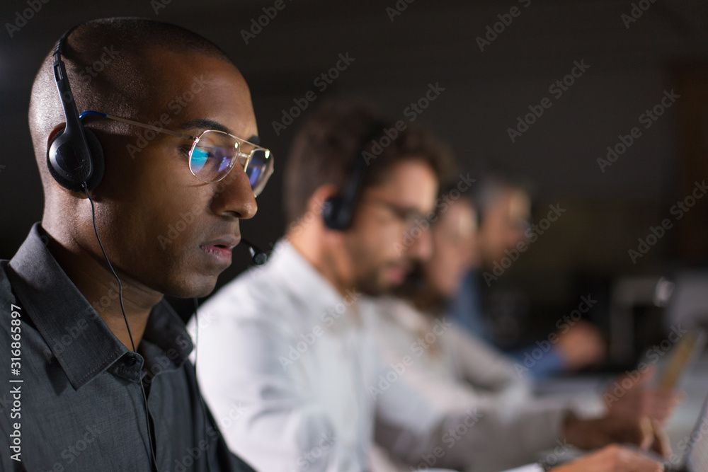 Concentrated call center operator communicating with client. African American young man in eyeglasses looking at laptop while serving client. Call center concept - obrazy, fototapety, plakaty 