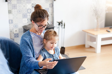 Mother and daughter using laptop and Internet. Freelancer workplace home. Woman and child girl sitting on sofa together. Concept of female business, working mom, freelance. Lifestyle moment.