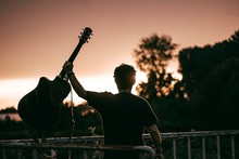 Man Holding A Guitar Near Metallic Fences With Greenery On The Blurry Background During Sunset