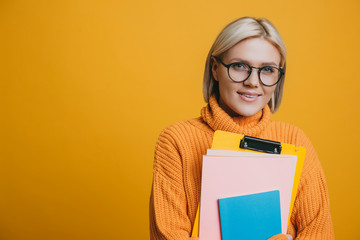 Portrait of a beautiful young blonde female student dressed in yellow looking at camera smiling while holding her books against yellow background.