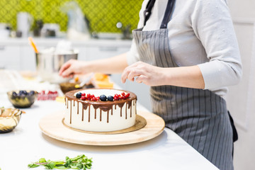 Wall Mural - Confectioner decorates with berries a biscuit cake with white cream and chocolate. Cake stands on a wooden stand on a white table. The concept of homemade pastry, cooking cakes.