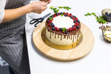 Confectioner decorates with berries a biscuit cake with white cream and chocolate. Cake stands on a wooden stand on a white table. The concept of homemade pastry, cooking cakes.