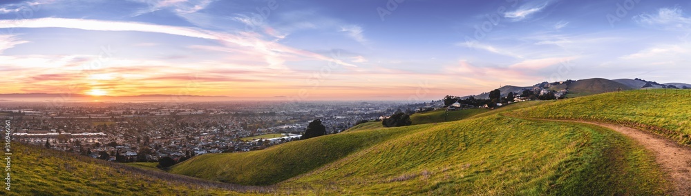 Sunset view of hiking trail on the verdant hills of East San Francisco Bay Area; the city of Hayward and the bay visible in the valley; California - obrazy, fototapety, plakaty 