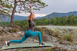 Blond pretty woman in green leggings with sexy tight booty is doing stretching by raising her hands and leaning against the mat in the picturesque place in mountains near the river and tree on stones.