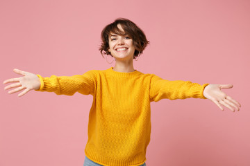 Wall Mural - Smiling young brunette woman girl in yellow sweater posing isolated on pastel pink wall background studio portrait. People lifestyle concept. Mock up copy space. Standing with outstretched hands.