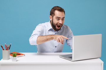 Wall Mural - Excited young bearded man in light shirt sit at desk isolated on pastel blue background. Achievement business career concept. Mock up copy space. Work on project, point finger on laptop pc computer.