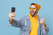 © ViDi Studio - Funny young hipster guy in fashion jeans denim clothes isolated on pastel blue background. People lifestyle concept. Mock up copy space. Doing selfie shot on mobile phone, showing thumb up, blinking.
