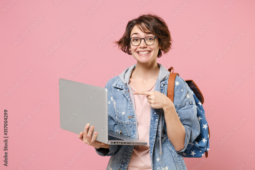 Smiling young woman student in denim clothes eyeglasses backpack isolated on pastel pink background. Education in high school university college concept. Pointing index finger on laptop pc computer. - obrazy, fototapety, plakaty 