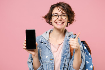 Smiling woman student in denim clothes glasses backpack isolated on pastel pink background. Education in school university college concept. Hold mobile phone with blank empty screen, showing thumb up.