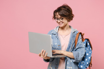 Smiling young woman student in denim clothes glasses backpack isolated on pastel pink background. Education in high school university college concept. Mock up copy space. Work on laptop pc computer.