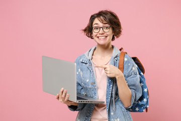 Smiling young woman student in denim clothes eyeglasses backpack isolated on pastel pink background. Education in high school university college concept. Pointing index finger on laptop pc computer.