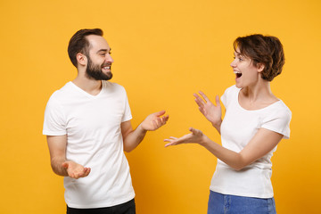 Funny young couple friends bearded guy girl in white t-shirts posing isolated on yellow orange background. People lifestyle concept. Mock up copy space. Looking at each other speaking spreading hands.