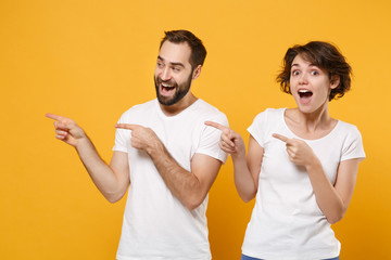 Surprised young couple friends bearded guy girl in white blank empty t-shirts posing isolated on yellow orange background. People lifestyle concept. Mock up copy space. Pointing index fingers aside.