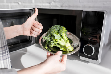 Wall Mural - cropped view of woman holding plate with broccoli near microwave and showing like