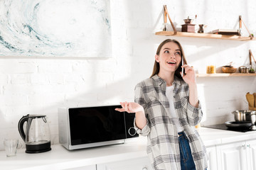 Wall Mural - smiling woman talking on smartphone near microwave in kitchen