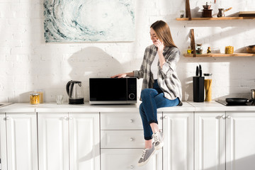 Wall Mural - smiling woman talking on smartphone near microwave in kitchen