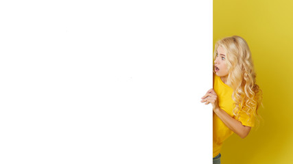 Wall Mural - Young happy woman peeks out from behind a white banner on a yellow background. Point to an empty blank on a form, a copy space for text. Horizontal shot