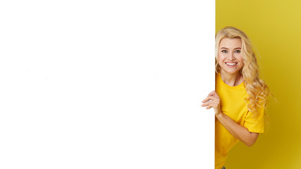Wall Mural - Young happy woman peeks out from behind a white banner on a yellow background. Point to an empty blank on a form, a copy space for text. Horizontal shot