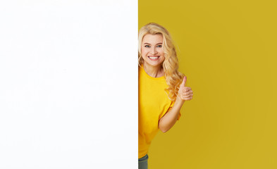 Wall Mural - Young happy woman peeks out from behind a white banner on a yellow background. Point to an empty blank on a form, a copy space for text. Horizontal shot