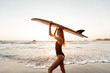 © Yevhenii - Surfer girl surfing looking at ocean beach sunset. Attractive young woman in black swimsuit is standing on beach with white surfboard in hands over her head