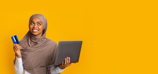 Doubtful afro muslim girl holding credit card and laptop in hands