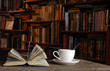 Open book. Nearby is a cup of coffee on a saucer and with a teaspoon. In the background shelves with books. Concept - coffee break, education, evening reading