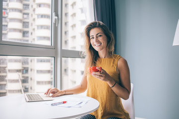 Confident female sitting at table with cup and documents tapping on notebook