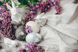 Fototapeta Kwiaty - Modern Easter eggs, white bunnies and lilac flowers on linen rural fabric. Stylish holiday table decor. Happy Easter. Space for text. Natural dyed easter eggs and spring flowers.