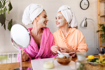 mother and daughter in bathrobes and towels on head using natural cosmetics and having fun together 