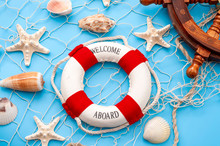 Maritime Shipping And Oceanic Sailing Conceptual Theme With Tabletop View Of Lifesaver, Fishing Net, Steering Helm And Marine Life (starfish And Shells) Isolated On Blue Background