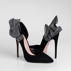 Womens black suede high heel shoes with a decorative bow element with shiny rhinestones on heel. Close-up side view. On a white background and natural shadows.