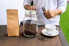 Person Grinding Coffee With Manual Coffee Grinder Beside Blur Of Blank Brown Paper Bag Foreground
