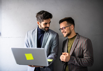 Two young businessman standing, one of them holding a laptop on a monochrome background.