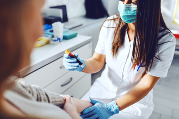 Female lab assistant in uniform, with protective mask and rubber gloves holding test tube with blood and holding patient's arm. Patient holding cotton on vein.