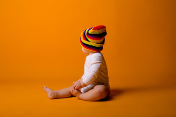 Wall Mural - baby boy in a white bodysuit and multicolored hat holding a toy, sitting on a yellow background, space for text