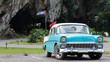 front of a parking baby-blue classic cuban car in a park with the cuban flag in  background