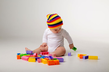 Wall Mural - baby boy in white bodysuit and multicolored hat playing with a multicolored plastic constructor on a white background isolate, child development concept, space for text