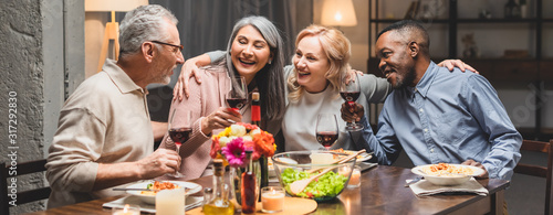 Fotografía panoramic shot of smiling multicultural friends hugging and holding wine glasses