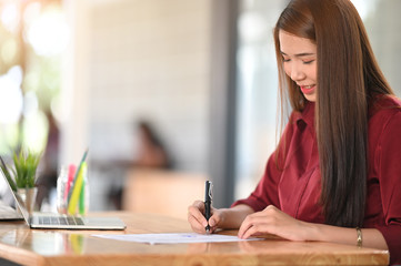 Wall Mural - Portrait of beautiful woman looking good in red shirt while writing on the paper and sitting at the wooden working desk.