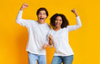 Overjoyed interracial couple celebrating success with raised fists over yellow background