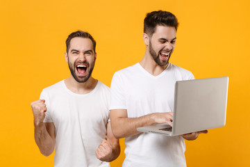 Sticker - Joyful young men guys friends in white t-shirts isolated on yellow orange background studio portrait. People lifestyle concept. Mock up copy space. Working on laptop pc computer doing winner gesture.