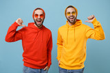 Fototapeta  - Two cheerful hipster men guys in fashion red yellow clothes eyeglasses posing isolated on pastel blue background in studio. People lifestyle concept. Mock up copy space. Pointing thumbs on themselves.